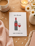 Card with wine bottle and glass illustration on a wooden table with coffee and flowers.