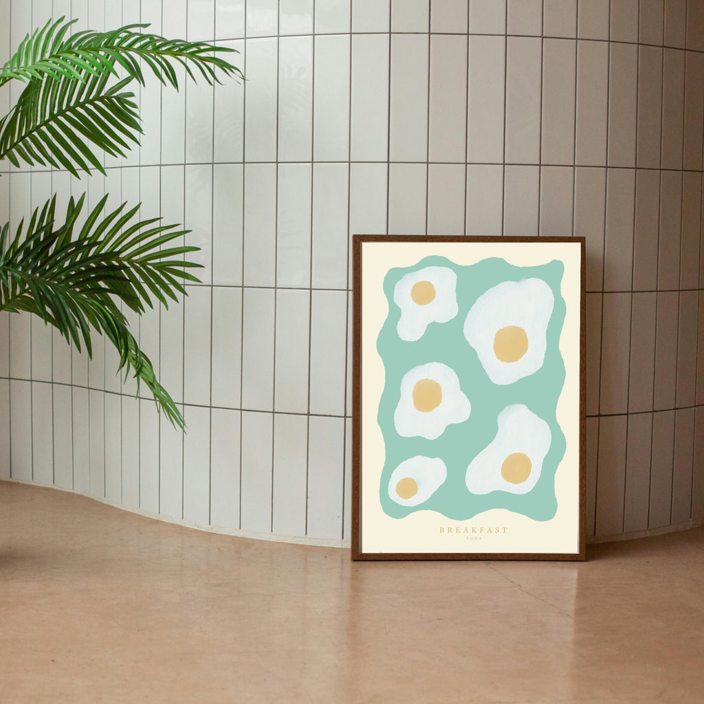 An A4 wall decor art print featuring a minimalist design with fried eggs on a teal background, placed against a tiled wall with a potted plant to the left.