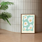 An A4 wall decor art print featuring a minimalist design with fried eggs on a teal background, placed against a tiled wall with a potted plant to the left.