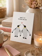 Card with penguin illustration and text on a table with books and a candle.