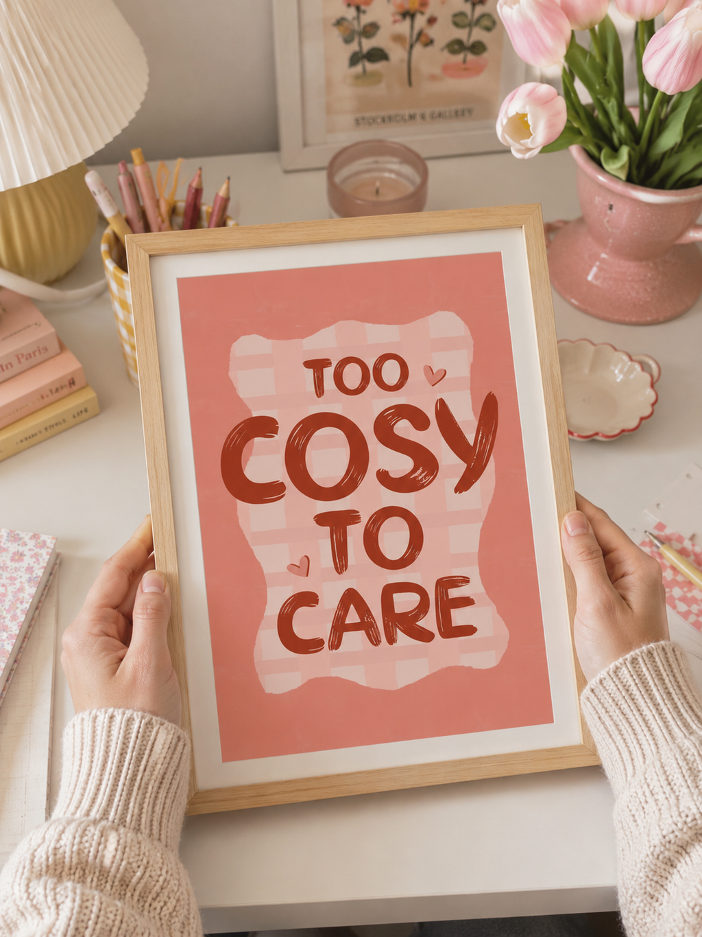 Framed print with 'Too Cosy To Care' text held by hands on a desk.