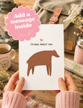 A person holds a white greeting card with a brown bear silhouette and the text 'I'M WILD ABOUT YOU' in a cozy, rustic setting. A pink cloud-shaped graphic overlay says 'Add a message inside'.
