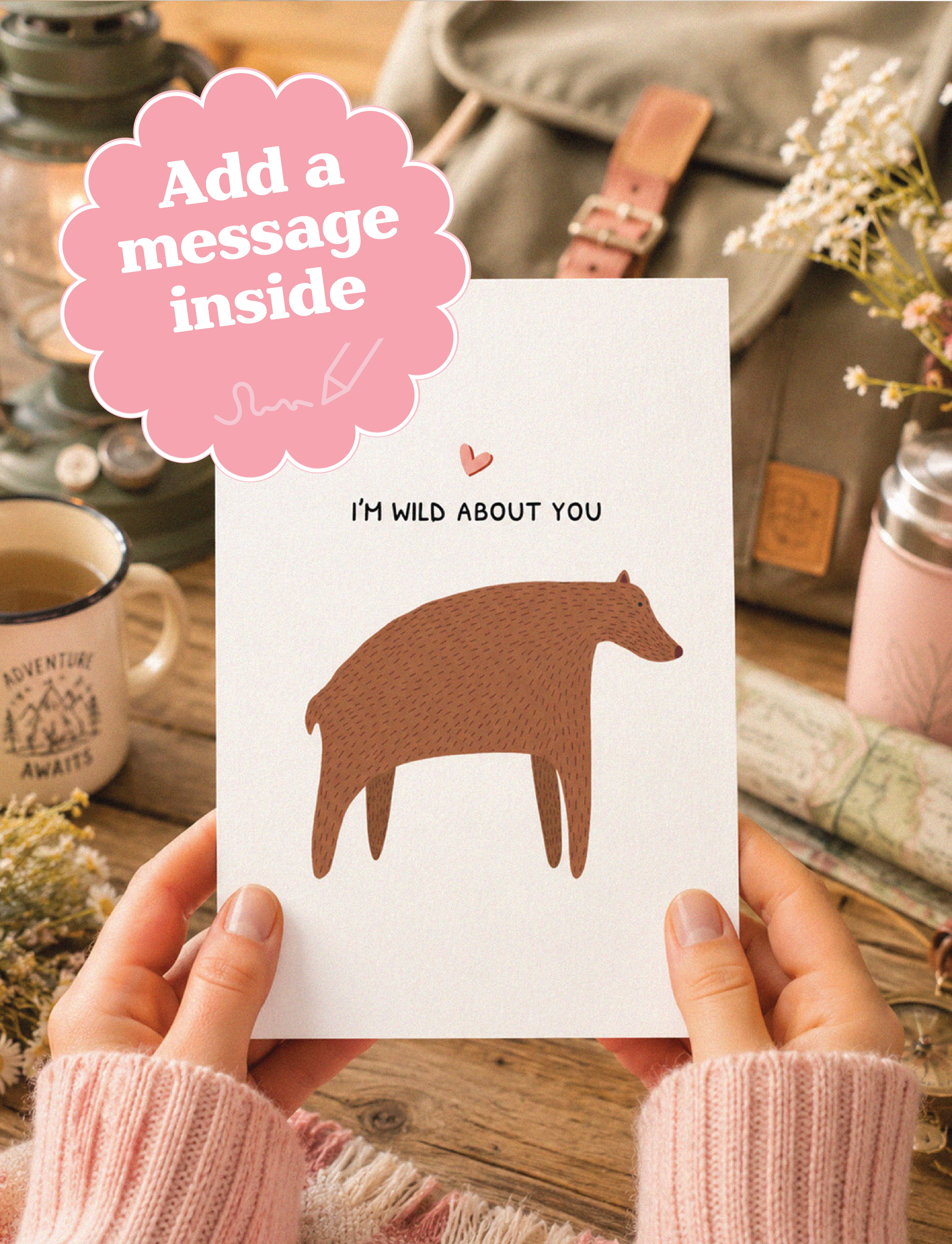 A person holds a white greeting card with a brown bear silhouette and the text 'I'M WILD ABOUT YOU' in a cozy, rustic setting. A pink cloud-shaped graphic overlay says 'Add a message inside'.